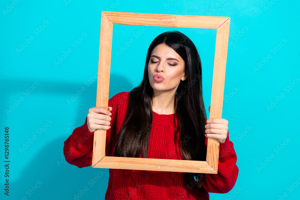 Stylish young woman in vibrant red sweater posing with wooden frame against turquoise background showing a fun and unique attitude Stylish young woman in vibrant red sweater posing with wooden frame against turquoise background showing a fun and unique attitude