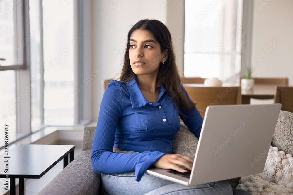Pensive young Indian female working on laptop seated on couch, looks into distance, search issue solution or creative ideas for online project, teleworking on-line at home using internet connection Pensive young Indian female working on laptop seated on couch, looks into distance, search issue solution or creative ideas for online project, teleworking on-line at home using internet connection