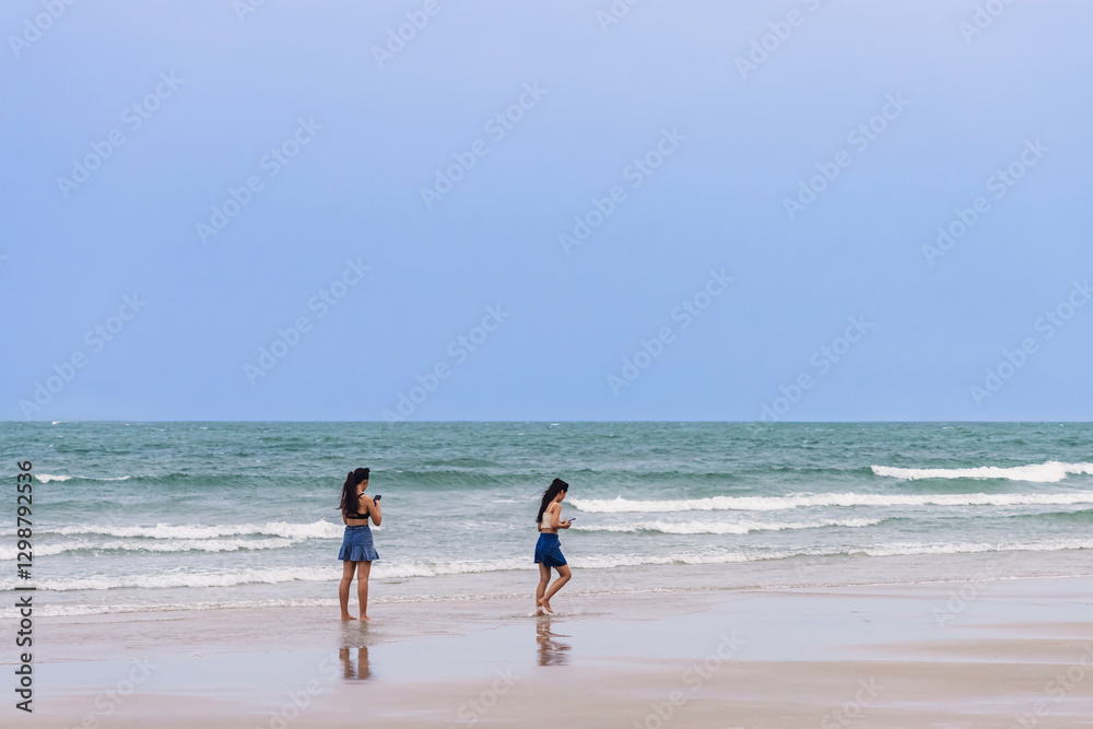 Back view of happy carefree women using mobile phone at beach. Woman friends using smartphone at seaside. Beautiful lovely two women spending time at beach. Concept of travel, tourism and vacation. Back view of happy carefree women using mobile phone at beach. Woman friends using smartphone at seaside. Beautiful lovely two women spending time at beach. Concept of travel, tourism and vacation.