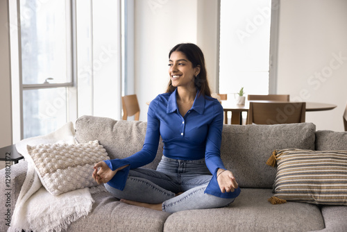 Young Indian woman sitting cross-legged on sofa in meditative pose in modern living room, practicing mindfulness or meditation, enjoy untroubled leisure alone at home. Inner, peace, balance