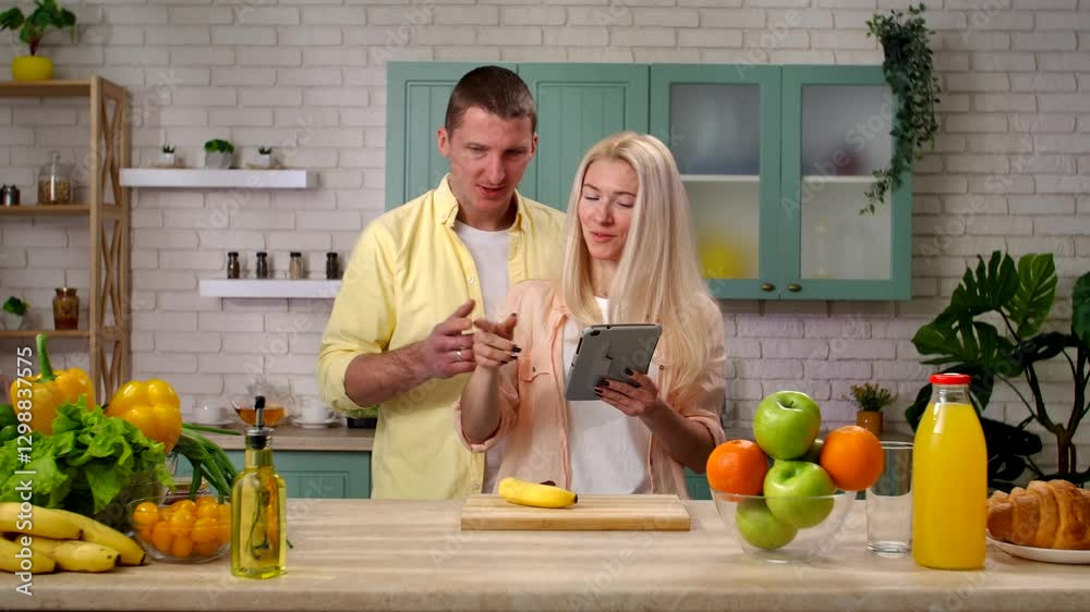 Couple of man and woman in the morning at home in the kitchen at table preparing breakfast, talking smiling, woman scrolling shows social media on tablet online. Slow motion. Couple of man and woman in the morning at home in the kitchen at table preparing breakfast, talking smiling, woman scrolling shows social media on tablet online. Slow motion.