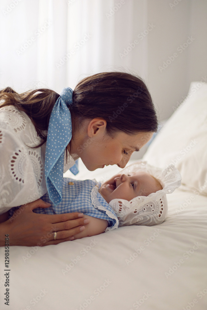 A beautiful brunette woman in a white dress with a baby in a cap lies on the bed at home light from the window.