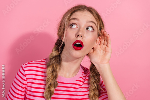 Фотография Young woman with blonde braids listening attentively on pink background, wearing stylish striped t-shirt and red lipstick