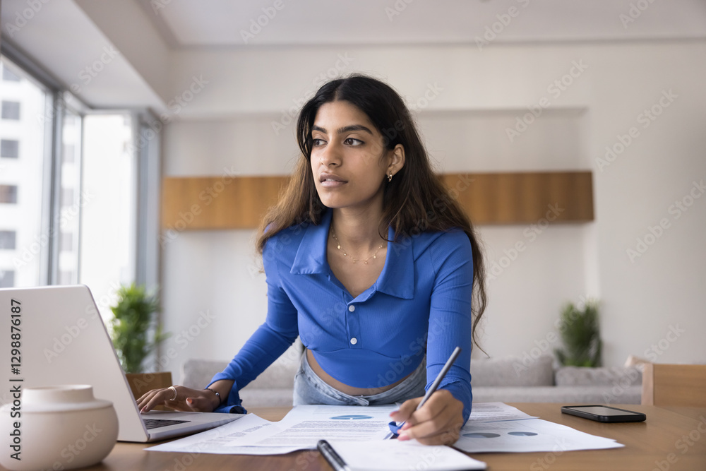 Serious focused Indian woman makes notes or corrections, analyze information, writing ideas or thoughts while working on project studying on coursework leaning at workplace desk with laptop and papers Serious focused Indian woman makes notes or corrections, analyze information, writing ideas or thoughts while working on project studying on coursework leaning at workplace desk with laptop and papers