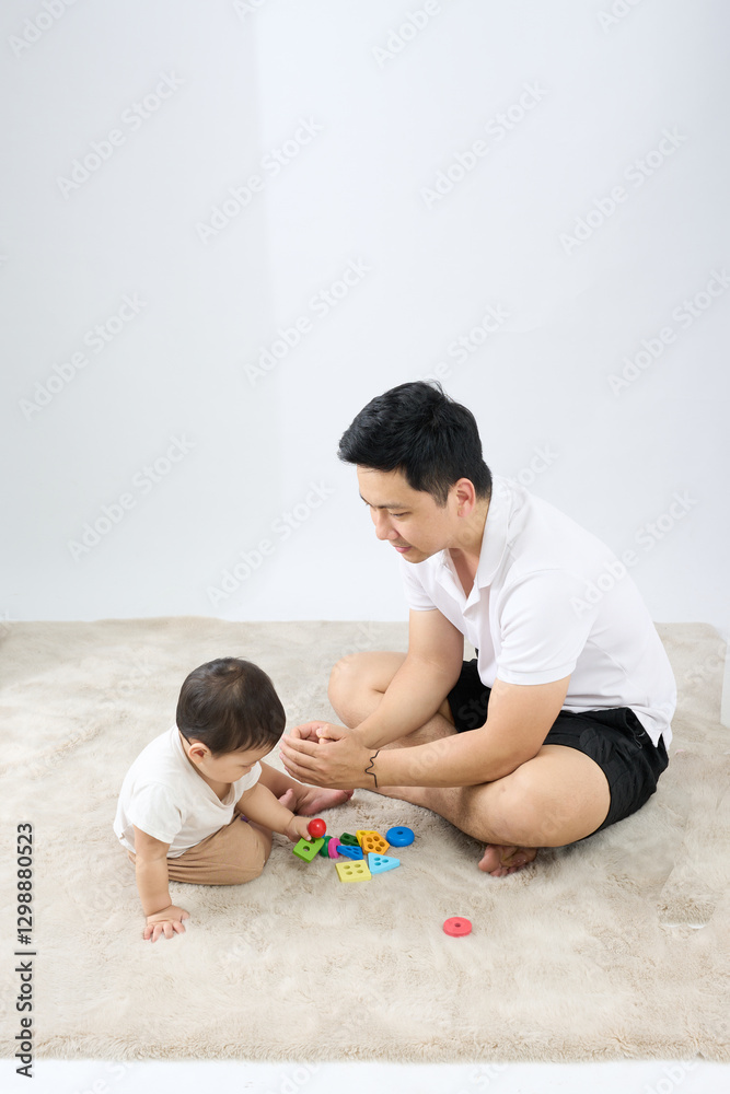 .The man is sitting on the floor with his baby and they are playing together. The man is holding a toy in front of the baby while the baby reaches out for it. .The man is sitting on the floor with his baby and they are playing together. The man is holding a toy in front of the baby while the baby reaches out for it.