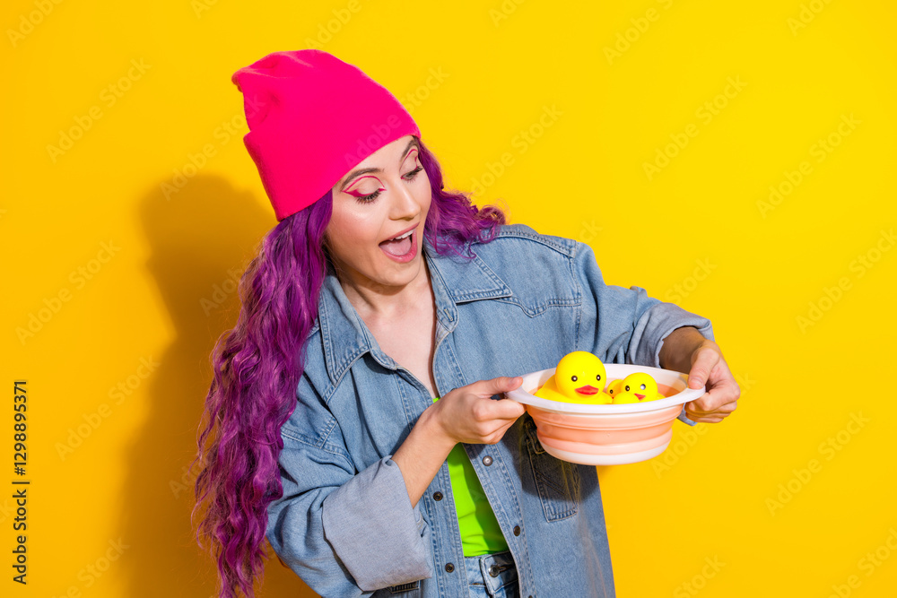 Young stylish woman with purple hair wearing a pink beanie and denim jacket holding rubber ducks on a yellow background Young stylish woman with purple hair wearing a pink beanie and denim jacket holding rubber ducks on a yellow background