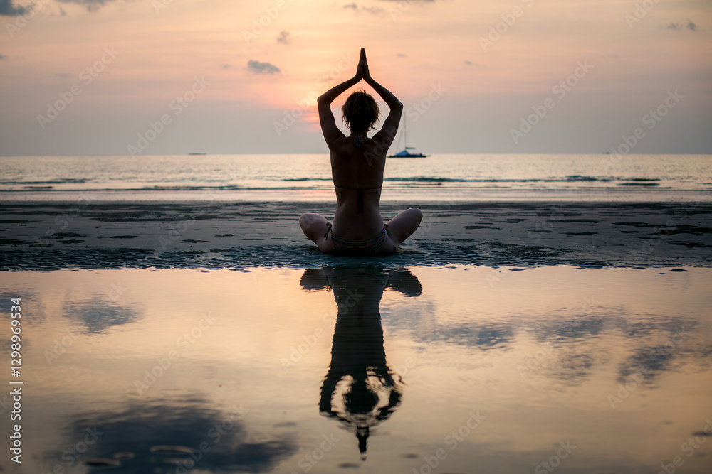 The silhouette of a yoga woman practicing on the beach with her reflection visible in the water.