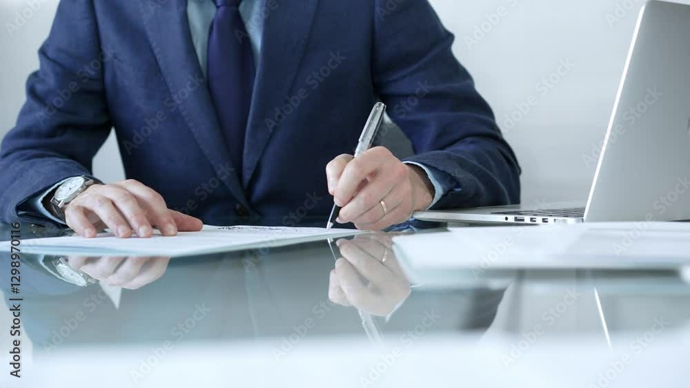 Businessman writing signature on contract with laptop and documents on glass desk in office. Business people concept