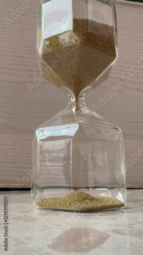 Glass hourglass with golden sand flowing slowly, closeup shot on reflective surface