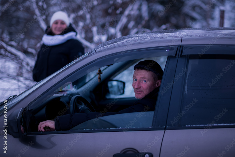 A man peeks out of his car on a snowy winter day, with a woman in the background.
