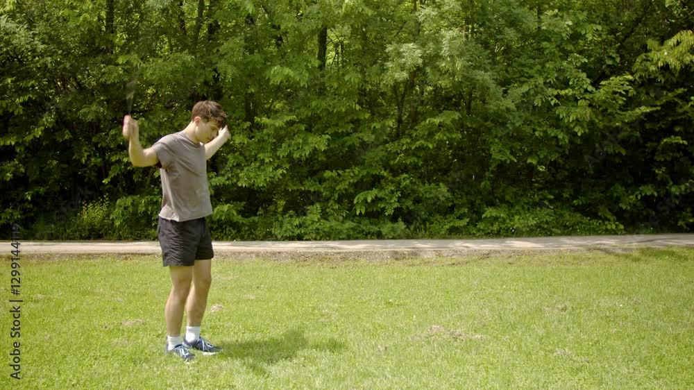 A young man exercises with a jump rope in a park on a sunny summer day. He is dressed in a gray t-shirt, black shorts, white socks, and blue sneakers. The background includes grass, trees, and a A young man exercises with a jump rope in a park on a sunny summer day. He is dressed in a gray t-shirt, black shorts, white socks, and blue sneakers. The background includes grass, trees, and a