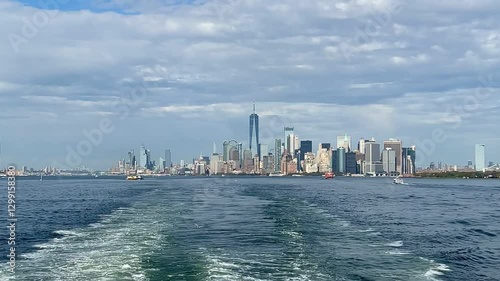 A breathtaking panoramic view of the Manhattan skyline as the Staten Island Ferry moves away. The wake contrasts with the city’s skyscrapers under a cloudy, blue sky.
