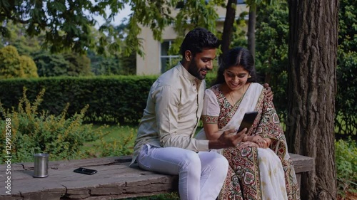 An Indian couple in traditional attire share a tender moment while looking at a phone. The outdoor setting with lush greenery adds a romantic touch
