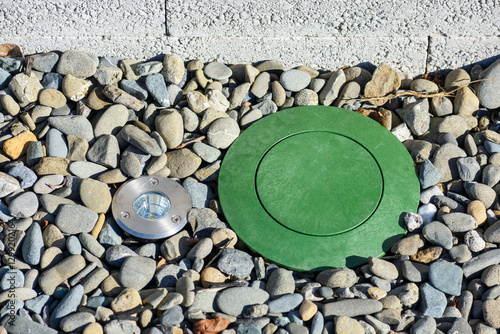 Green pop-up drainage emitter installed on a pebble-covered surface, accompanied by a circular stainless steel ground light