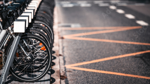 Fototapeta Naklejka Na Ścianę i Meble -  Row of parked rental bicycles aligned at a docking station near a road with orange markings. Close-up of front wheels and spokes in sharp focus, background blurred. Copy space area on the right