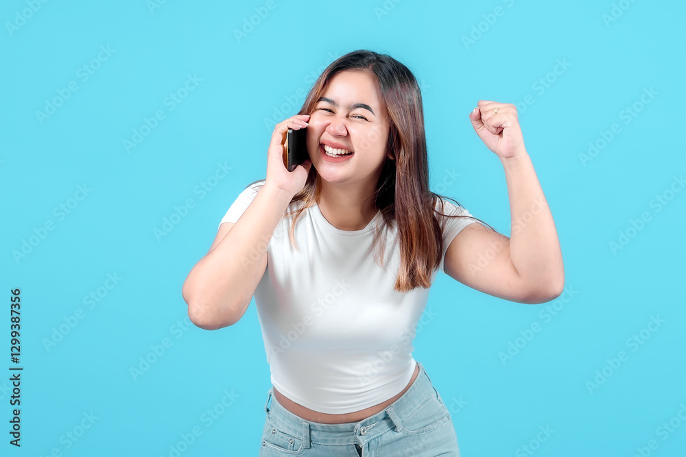 Fototapeta premium A cheerful young woman celebrates while talking on her smartphone, expressing joy and excitement against a bright blue background