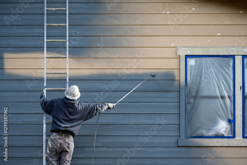 Fototapeta Naklejka Na Ścianę i Meble -  Professional painter in hard hat and air respirator on extension ladder with paint sprayer painting back exterior of house, job site for new housing development
