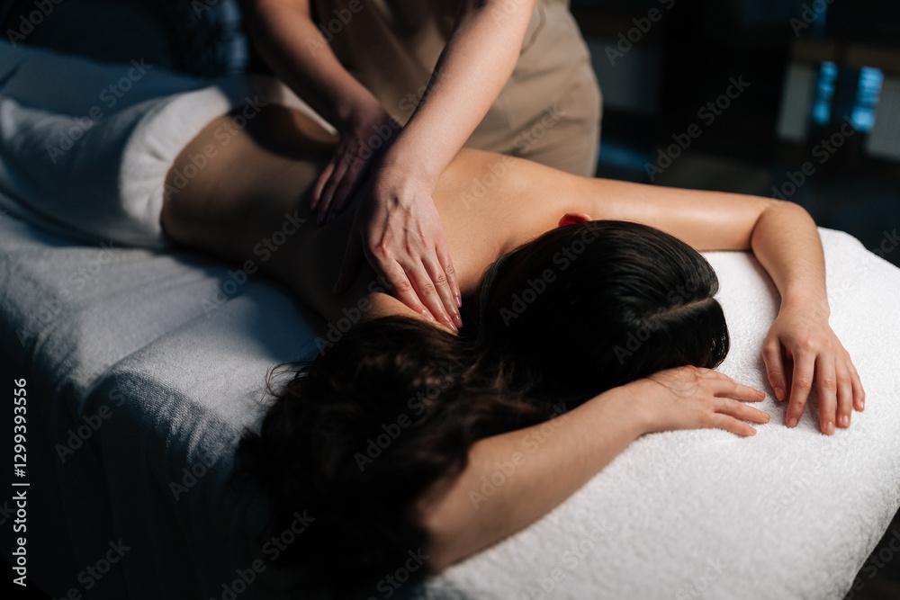 Obraz premium Cropped shot of skilled massage therapist working on female client shoulders muscles during therapeutic session, providing deep tissue relaxation in serene, softly illuminated wellness environment.