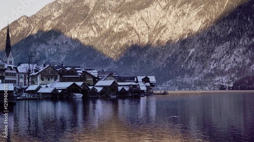 Picturesque Hallstatt alpine village with snow-covered rooftops sits by a tranquil lake, surrounded by towering mountains in a serene winter setting.
