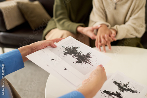 Female therapist holding inkblot test in hands with teenage girl and mother sitting together holding hands in background, mental health assessment session, copy space.