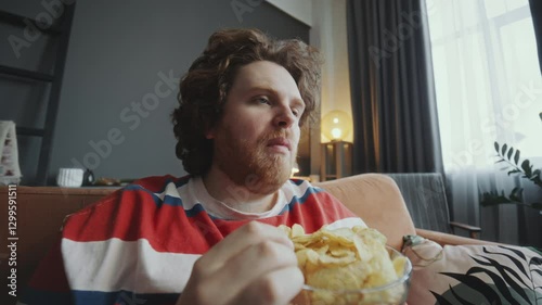 Young obese man relaxing on couch, eating potato chips from bowl and watching TV during day at home. Whip zoom shot