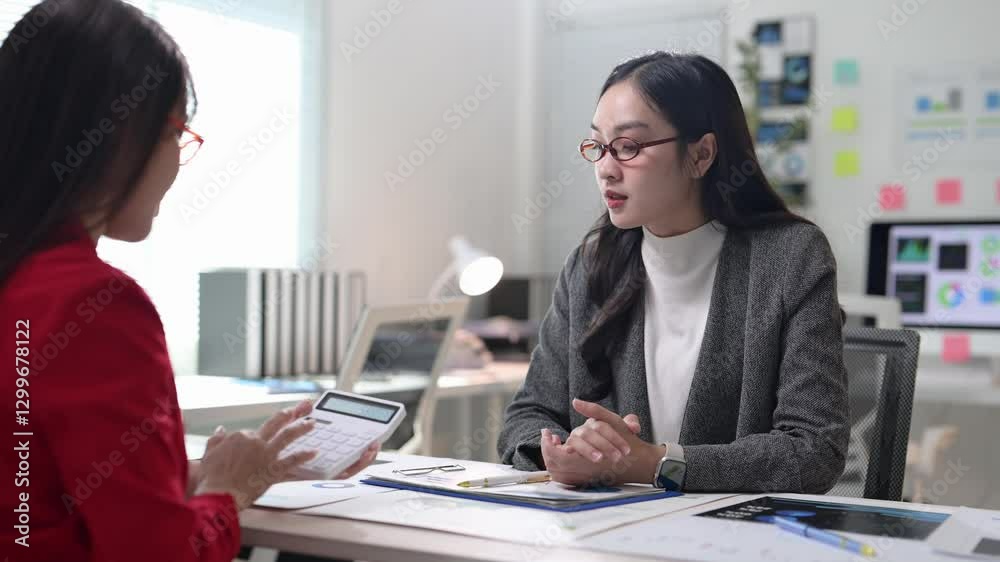 A woman is sitting at a desk with a calculator and a piece of paper. She is talking to another woman A woman is sitting at a desk with a calculator and a piece of paper. She is talking to another woman