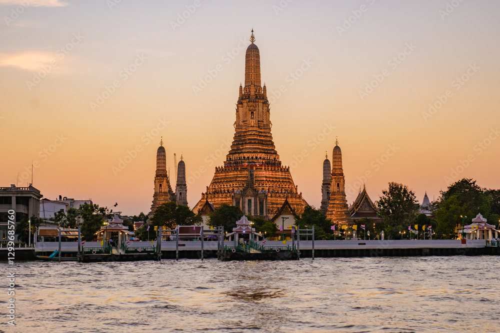 Naklejka premium As the sun sets over Bangkok, Wat Arun stands majestically by the river, bathed in golden light. Visitors admire the stunning architecture and vibrant colors, creating a serene atmosphere at dusk.