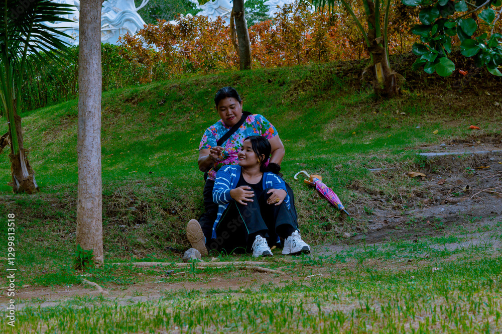 Fototapeta premium A mother and her teenage girl are sitting casually under a tree in a serene garden park, enjoying a cheerful conversation, surrounded by lush greenery.