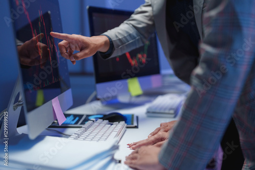 Two stockbrokers are examining financial charts displayed on computer screens, analyzing market trends and investment opportunities in a dimly lit office, working late into the night