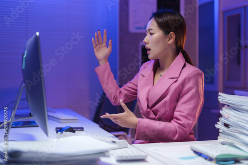 Young asian businesswoman in a pink suit experiencing stress during a late-night video call in the office, arguing and gesturing with her hands while managing overwhelming pressure