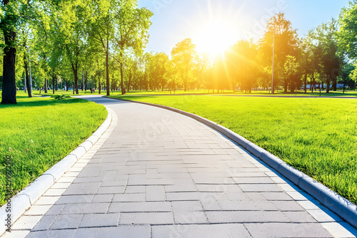 Sunny park pathway, paved walkway curving through vibrant green grass and lush trees, idyllic summer scene.