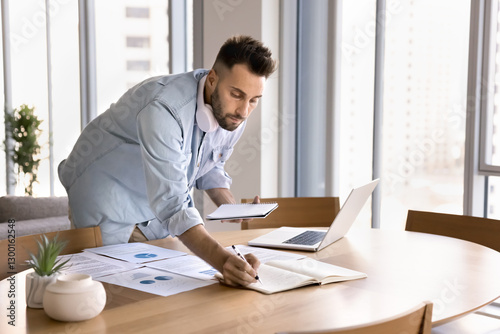 Focused man multitasking, working on analytical task, prepare presentation or conducting research standing at table, holds copybook while writes, laptop, printed documents spread across workplace desk