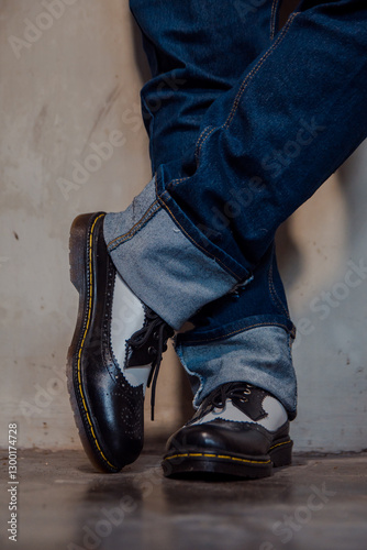 Close up of a man's legs wearing blue jeans paired with two-tone black and white genuine leather brogue shoes. Close up of shiny vintage brogue shoes against a dirty white wall.