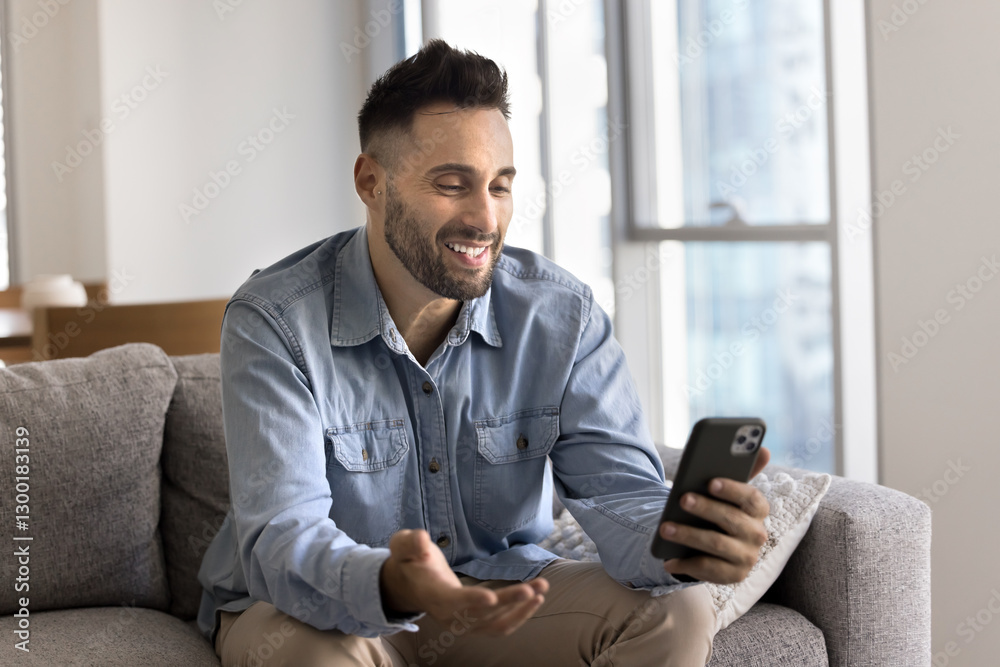 Latin man seated comfortably on sofa, holding smartphone engaging in lively videocall digital conversation using mobile application, smile while get pleasant news, receive updates, enjoy new software Latin man seated comfortably on sofa, holding smartphone engaging in lively videocall digital conversation using mobile application, smile while get pleasant news, receive updates, enjoy new software