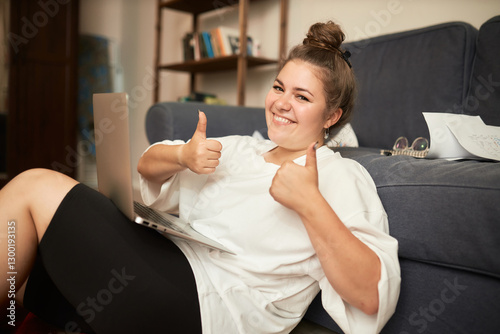 Happy woman giving excellent mark for software or food delivery application, having rest ordering dinner online sitting on floor next to sofa holding laptop on laps and showing thumbs up