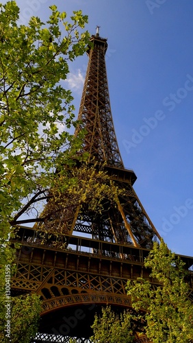 Visitors stand beneath the towering Eiffel Tower in Paris, marveling at its intricate design. Green trees frame the iconic structure under a clear blue sky, perfect for exploration