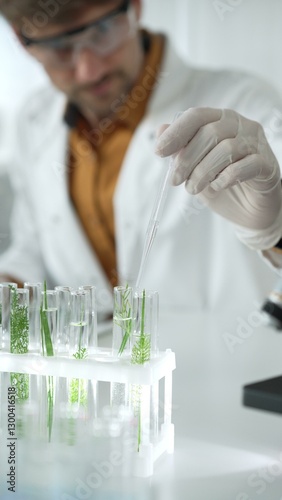Man scientist wearing a lab coat, white gloves and protective glasses, is dripping liquid from a pipette into a test tube with a green plant inside, vertical close up view. Science and medicine