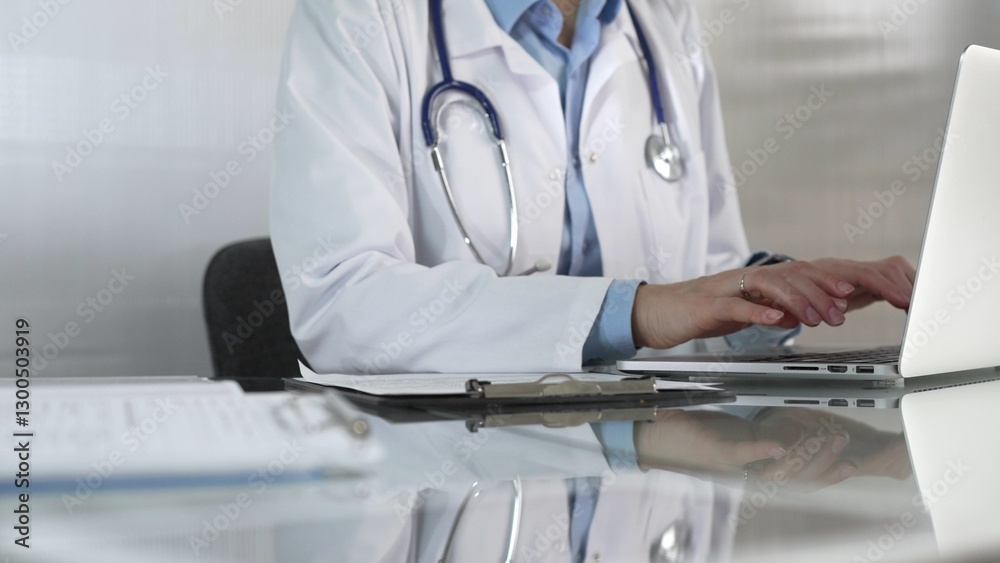 Doctor woman, wearing a blue blouse under a medical coat, is using technology in healthcare, close up of hands typing on laptop with stethoscope, glasses, and clipboard on desk. Medicine concept Doctor woman, wearing a blue blouse under a medical coat, is using technology in healthcare, close up of hands typing on laptop with stethoscope, glasses, and clipboard on desk. Medicine concept