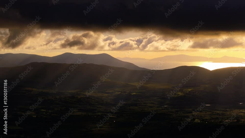 Dark clouds partially obscure the setting sun. Golden light streams through gaps, illuminating the distant mountains and valleys below. Landscape features are clearly visible. Dark clouds partially obscure the setting sun. Golden light streams through gaps, illuminating the distant mountains and valleys below. Landscape features are clearly visible.