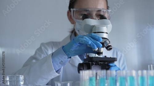 Scientist woman, wearing a lab coat, mask, blue gloves and safety glasses, is adjusting a microscope surrounded by test tubes in laboratory. Medicine and science concept