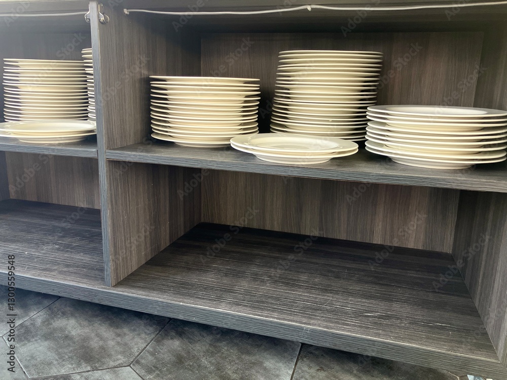 A pile of several clean white dinner plates in the bottom drawer of a buffet-style dining table, neatly arranged high A pile of several clean white dinner plates in the bottom drawer of a buffet-style dining table, neatly arranged high