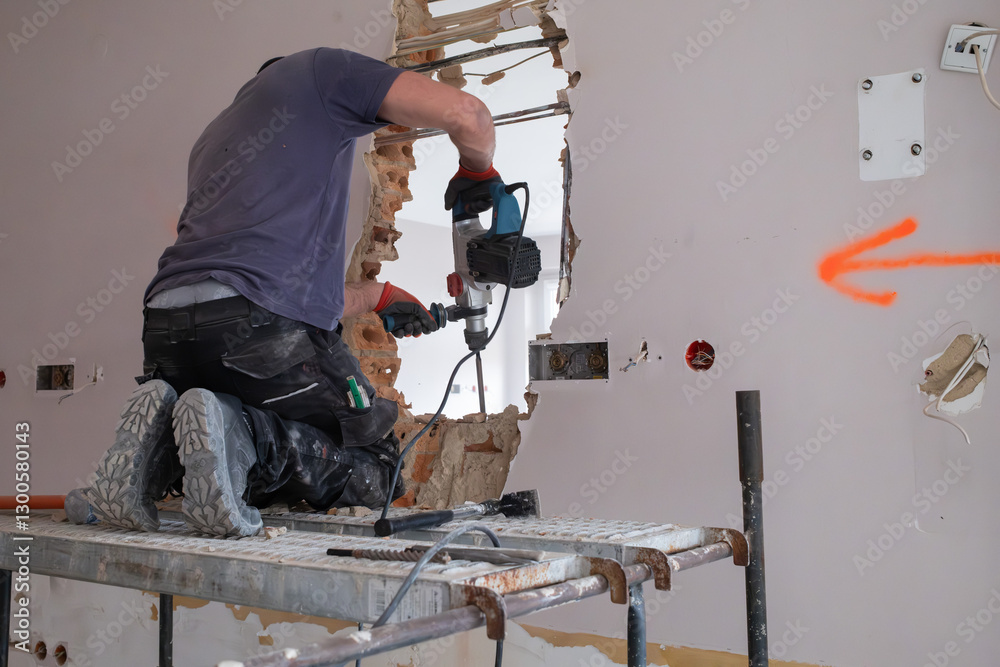 A construction worker on scaffolding demolishes a wall in a building with a hammer drill.