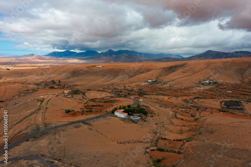 Aerial view of arid Fuerteventura landscape with isolated rural house surrounded by dramatic mountains. Barren desert terrain under stormy clouds with winding roads and sparse vegetation in the Canary