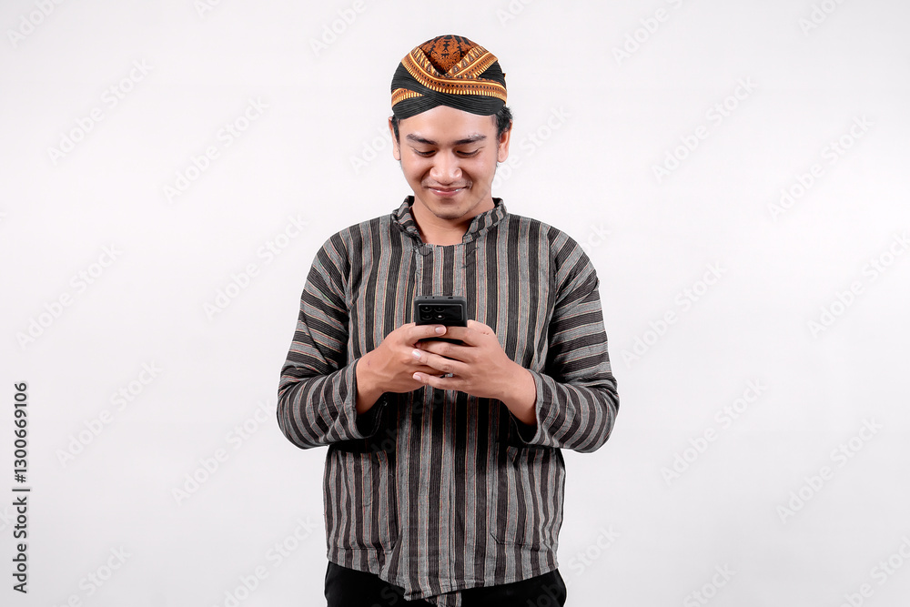 A young Indonesian man dressed in traditional sorjan or lurik attire smiles while looking at his smartphone, showcasing a blend of cultural heritage and modern technology against a neutral background. A young Indonesian man dressed in traditional sorjan or lurik attire smiles while looking at his smartphone, showcasing a blend of cultural heritage and modern technology against a neutral background.