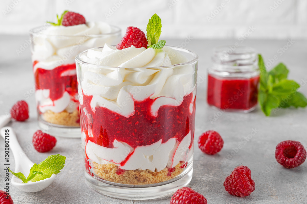 Raspberry trifle with whipped cream, berry sauce, cookie crumbs and mint leaves in a glass on a gray concrete background. Summer diet dessert. Raspberry trifle with whipped cream, berry sauce, cookie crumbs and mint leaves in a glass on a gray concrete background. Summer diet dessert.