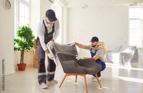 Two professional movers in uniform carefully unpacking and setting up armchair in bright spacious room. Loaders providing relocation service, handling furniture and organizing home moving process.