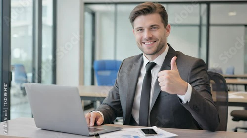 Professional man in suit giving thumbs up at office desk with laptop for business success
