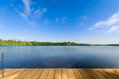 A summer day with a calm blue lake and a wooden dock in the foreground in Algonquin Provincial Park, Ontario, Canada. The water reflects the clear sky, surrounded by lush green trees along the shore.
