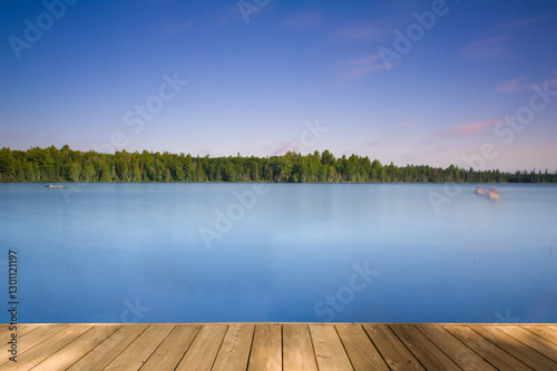 A long-exposure shot of a calm lake with a wooden dock in the foreground in Algonquin Provincial Park, Ontario, Canada. The water shows soft canoe movements. Sunset casts orange tones in the blue sky.