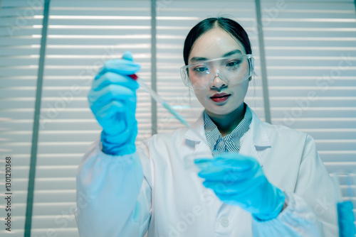 Woman in a lab coat is holding a beaker of blue liquid. She is wearing gloves and a lab coat